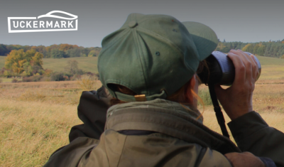 Eine Person von hinten blick tmit einem Fernglas in die Landschaft. Daneben das Logo der Regionalmarke Uckermark